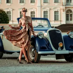 Elegancka kobieta w kwiecistej sukience retro z lat 20. pozuje przy zabytkowym kabriolecie przed pałacem.
