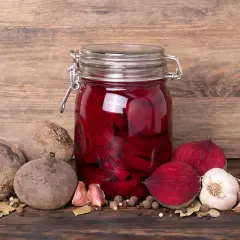 Ein Glas mit eingelegten, roten Bete Scheiben steht auf einem Holztisch. Daneben liegen frische rote Bete, Knoblauch und Gewürze. Das Bild zeigt den Prozess des rote Bete fermentieren.