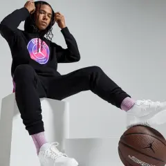 Mężczyzna w czarnej bluzie PSG Jordan z logo, czarnych spodniach dresowych, białych butach i różowych skarpetkach siedzi na białych sześcianach.