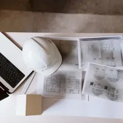 Na biurku leży laptop, biały kask, plany architektoniczne i poziomica. To kompletny zestaw do pracy nad projektem technicznym i prawem budowlanym.