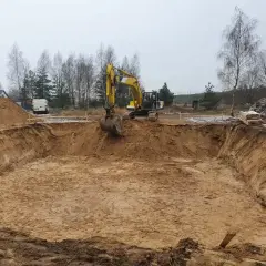 Żółta koparka pracuje przy wykopy pod fundamenty. W tle drzewa i biały van.