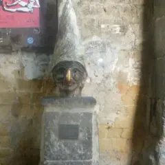 Busto con cappello a cono e un buffo naso di Pulcinella, su un piedistallo di pietra.