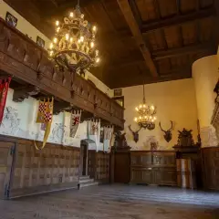 Wnętrza Zamku Czocha: bogato zdobiona sala z drewnianymi panelami, galerią, żyrandolami i trofeami łowieckimi.