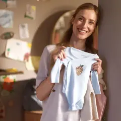 Glad mamma visar upp en ljusblå babybody med ett gulligt ekollonmotiv. Hon är veckans babe och ser fram emot att klä sin lilla skatt.