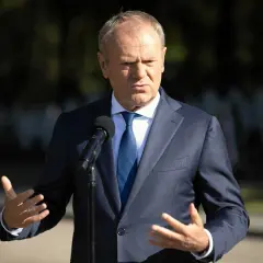 Donald Tusk w garniturze przemawia przy mikrofonie, gestykulując rękami.
