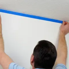 Mężczyzna przykleja niebieską taśmę malarską, by zamaskować krzywy sufit.