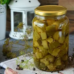 Słoiki pełne domowych pikli z ogórków, obok świeże ogórki, czosnek i koper.