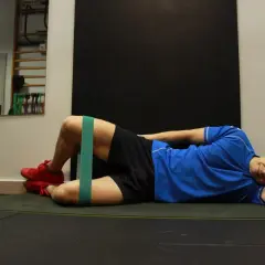 Mężczyzna wykonuje trening oporowy z gumą na udach, leżąc na macie. Ćwiczenie na pośladki.