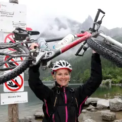 Uśmiechnięta rowerzystka z rowerem nad Morskim Okiem. W tle góry i tablica szlaku.