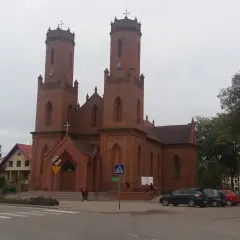 Imponujący kościół w Krokowej z cegły, z dwiema wieżami i zegarami, otoczony zielenią i samochodami.
