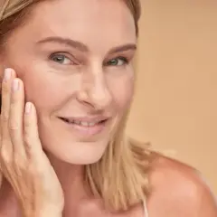 Uśmiechnięta kobieta z blond włosami dotyka policzka, prezentując naturalny wygląd skóry dzięki dobremu podkładowi kryjącemu bez efektu maski.