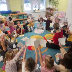 Dzieci w przedszkolu siedzą na dywanie i trzymają w górze rysunki z buźkami.