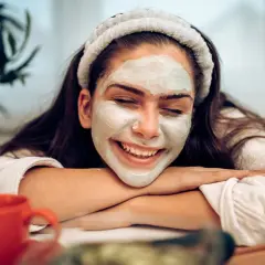 Lächelnde Frau mit Gesichtsmaske, die sich fragt: nach Gesichtsmaske eincremen oder nicht? Entspannter Moment mit rotem Becher.
