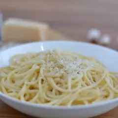 Ein Teller mit dampfenden Spaghetti, bestreut mit frisch geriebenem Parmesan. Dazu ein Stück Parmesan, eine Reibe und Knoblauch – perfekt für ein authentisches italienisches Gericht.