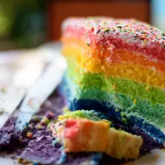 Ein Stück bunter **Regenbogen Kuchen** mit bunten Streuseln. Ein Stück ist bereits angeschnitten und liegt daneben.
