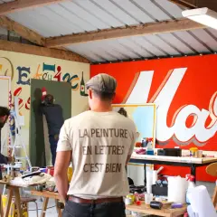 Des peintres travaillent dans un atelier, utilisant du matériel de peinture pour créer des lettres sur un mur. Le t-shirt d'un homme porte l'inscription "La peinture en lettres, c'est bien".