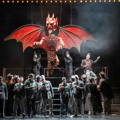 Bühnenszene der Oper Wuppertal: Eine Teufelsmarionette schwebt über Darstellern in Uniformen und Zylindern.