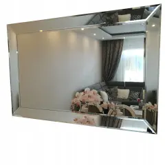 Eleganckie lustro fazowane w ramie, odbijające salon z kanapą i kwiatami.