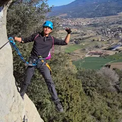 Un escalador sonriente, con casco azul, disfruta de la vía ferrata, un ejemplo de los diversos tipos de escalada, con un pueblo y campos verdes de fondo.