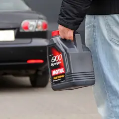Mężczyzna niesie kanister z olejem silnikowym Motul 6100 Synergie+ SAE 10W40. Za nim stoi czarny samochód.