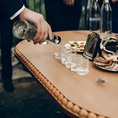 Na stole stoją kieliszki, a mężczyzna nalewa do nich wódkę. Obok wędliny, ciasta i kanapki. Zastanawiasz się, ile wódki na osobę?