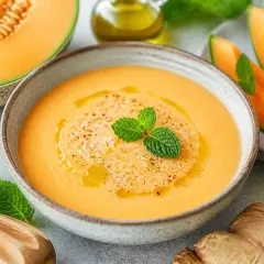 Délicieuse soupe de melon rafraîchissante, garnie de menthe fraîche et d'un filet d'huile d'olive. Des tranches de melon, du gingembre et une moitié de citron vert complètent cette scène estivale.