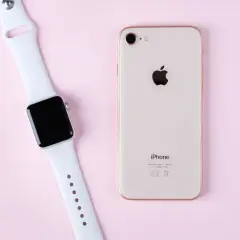 Apple Watch und iPhone liegen nebeneinander auf rosa Untergrund, bereit zum Laden.