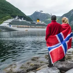 Para podróżuje po Norwegii, z flagą narodową i statkiem wycieczkowym w tle. Planujesz podróż? Pomyśl o ubezpieczenie Norwegia.