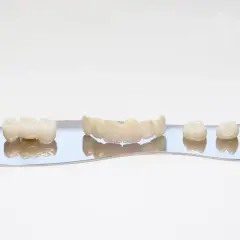 Zęby ceramiczne, w tym mostek i pojedyncze korony, prezentowane na lustrzanej powierzchni. Wykonane z materiałów takich jak korona cyrkonowa, zapewniają estetykę i trwałość.