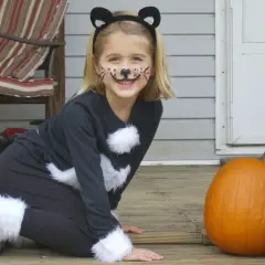 Bambina con trucco da gatto e orecchie, pronta per vestiti carnevale veloci fai da te bambini. Zucca arancione accanto.