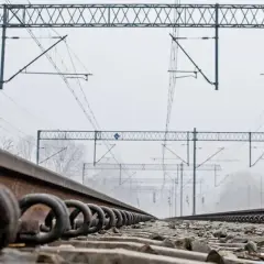 Zbliżenie na torowisko kolejowe z widocznymi kółkami mocującymi szyny. W tle sieć trakcyjna i mgliste drzewa.