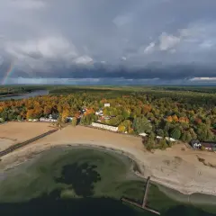 Jesioro Skorzęcin jesienią. Tęczowy łuk nad ciemnymi chmurami, piaszczysta plaża i kolorowy las.