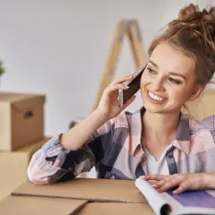 Uśmiechnięta kobieta rozmawia przez telefon, otoczona kartonami. Dowiaduje się, jak się zameldować w nowym miejscu.