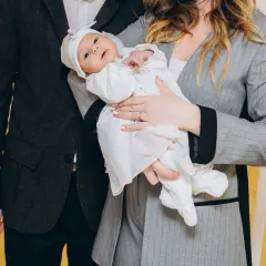 Elegancko ubrani rodzice trzymają niemowlę w białej szatce. Idealna stylizacja, jak ubrać się na chrzest wnuczki.