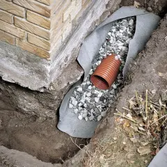 Drainage für ein Haus: Ein Drainagerohr liegt in Kies, umgeben von Vlies, neben dem Fundament.