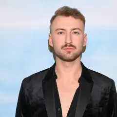 Michał Bartkiewicz w czarnym aksamitnym marynarkowym stroju, z zarostem i fryzurą, na tle błękitnego nieba. Jego życie prywatne budzi zainteresowanie.