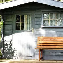 Ein kleines Gartenhaus mit Holzbänken davor. Das Dach ist mit Dachpappe abgedichtet, um es vor Regen zu schützen.