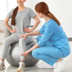 Une kinésithérapeute aide une patiente assise sur un ballon de gym à retrouver de la force dans les jambes grâce à des exercices ciblés.