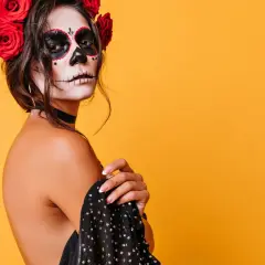 Kobieta z makijażem sugar skull i czerwonymi różami we włosach na pomarańczowym tle.