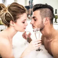 Para zbliża się do siebie w wannie pełnej piany, trzymając kieliszki szampana. Intymny moment, który zapowiada seks w wannie.