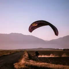 Parapentista aterrizando en un campo polvoriento al atardecer, rodeado de montañas. Una escena de turismo activo.