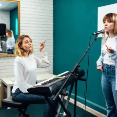 Lekcja śpiewu w domu: nauczycielka przy keyboardzie instruuje uczennicę przed mikrofonem.