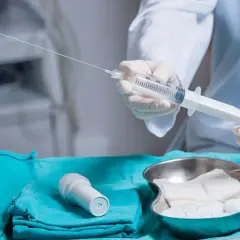 Lekarz przygotowuje się do amniopunkcji, trzymając strzykawkę z długą igłą. Na stole leżą też gaziki i inne akcesoria medyczne.