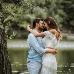 Para młoda, w białej sukni i błękitnej koszuli, przytula się czule nad jeziorem. Ich uśmiechy i spojrzenia sugerują szczęśliwą noc poślubną.