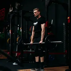 Ein Mann mit Bart und schwarzem T-Shirt hebt eine Langhantel im Fitnessstudio. Er trainiert seine Handkraft, um stärker zu werden.