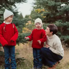 Urocza sesja zdjęciowa dla dziecka w otoczeniu choinek. Mama i dwie córki w czerwonych sweterkach i białych czapkach cieszą się wspólnym czasem.