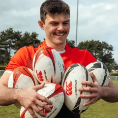 Uśmiechnięty mężczyzna w czerwonej koszulce trzyma kilka piłek do rugby i futbolu amerykańskiego.