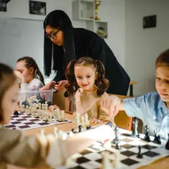 Nauczycielka pokazuje dzieciom, jak grac w szachy. Uczniowie z uwagą obserwują ruchy figur na szachownicy.
