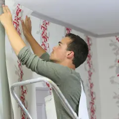 Mann auf Leiter tapeziert Wand mit rotem Blumenmuster. So tapeziert man Schritt für Schritt.