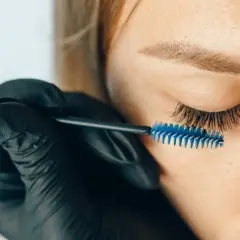 Stylist w czarnej rękawiczce szczotkuje przedłużone rzęsy. Zastanawiasz się, jak długo odrastają rzęsy po przedłużaniu?